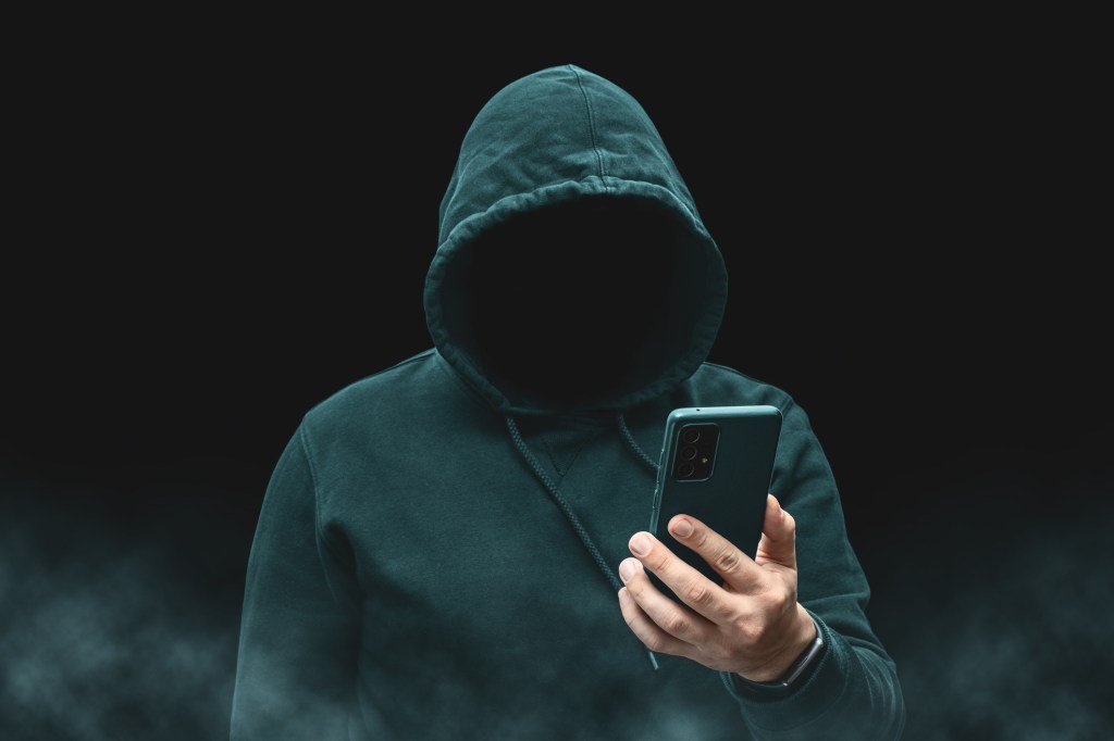 The silhouette of a faceless, hooded figure apparently a hacker or criminal online, holding a smartphone